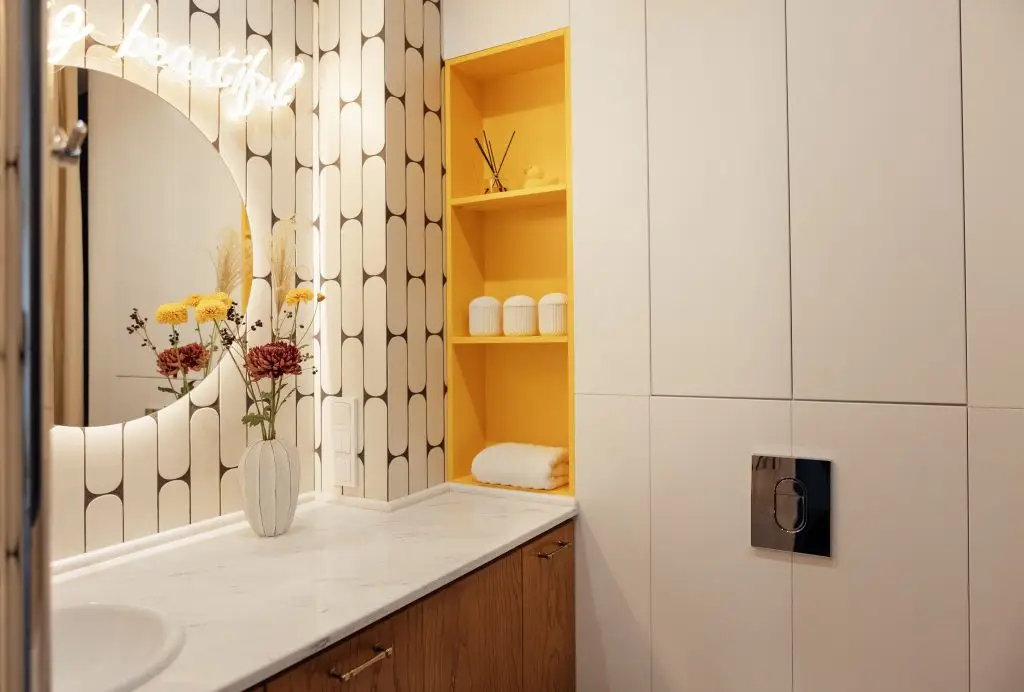 Modern stylish bathroom interior. A marble countertop with a sink and a vase. A round illuminated mirror. Bright accent on the yellow shelves with household things.